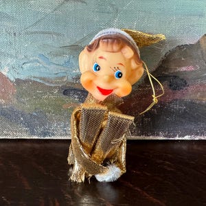 May include: A vintage Christmas elf ornament with a smiling face, blue eyes, and a red mouth. The elf wears a gold and brown outfit with a white pom-pom and a gold hat. The background is a painting with blue, brown, and green tones.