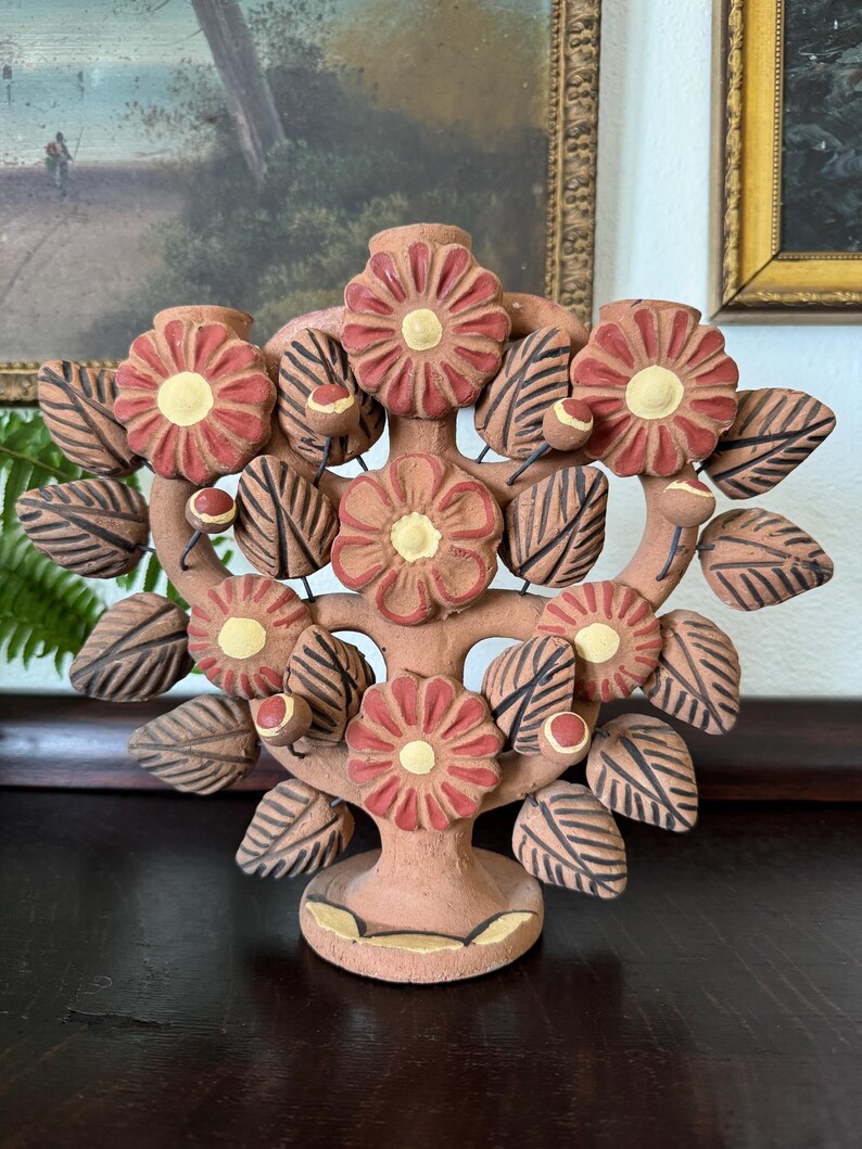 Pu&ograve; includere: Un candelabro in terracotta a forma di albero con cinque portacandele. L'oggetto presenta fiori rossi scolpiti con centri gialli e foglie a strisce marrone scuro. La base &egrave; circolare con accenti gialli e sono presenti piccoli boccioli.