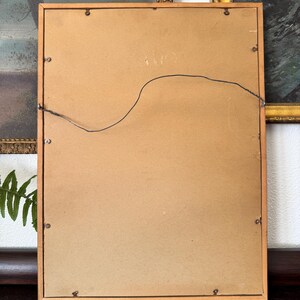 May include: The back of a wooden picture frame with a brown cardboard backing and a wire hanger attached. The frame has a light wood finish.