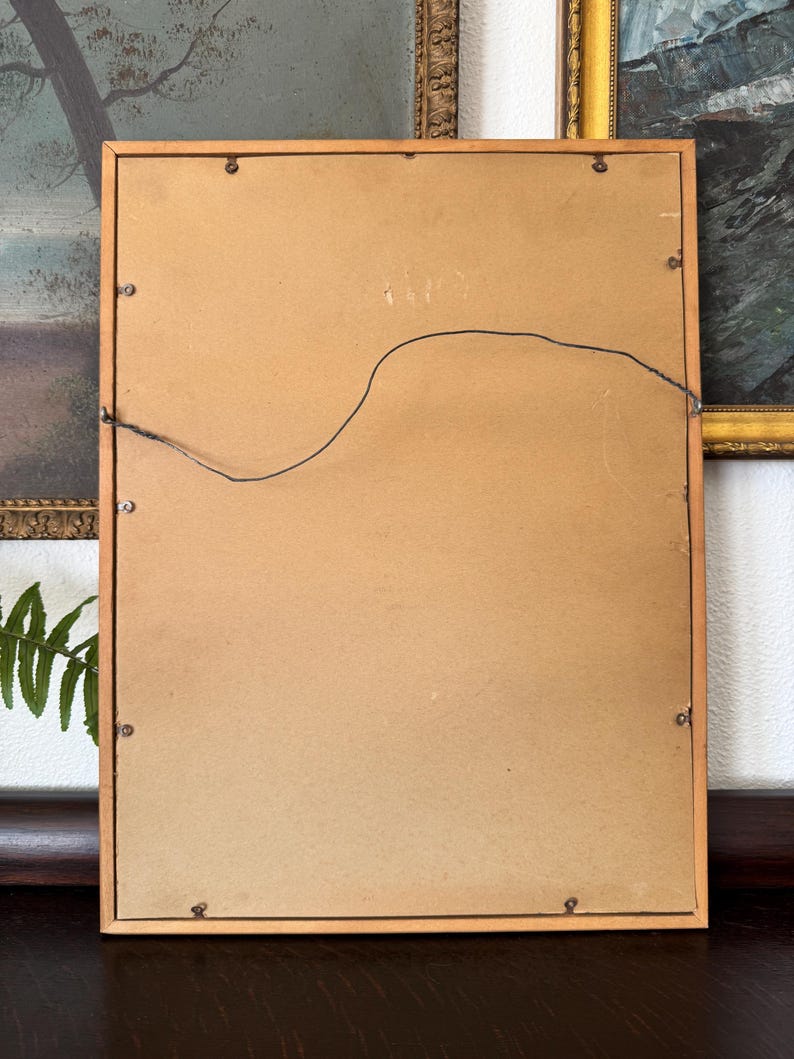 May include: The back of a wooden picture frame with a brown cardboard backing and a wire hanger attached. The frame has a light wood finish.