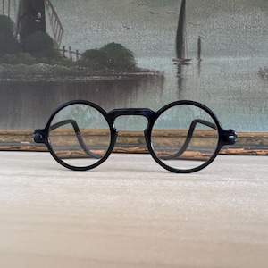May include: A pair of black round-frame glasses with clear lenses. The frames feature a circular design with a bridge connecting the two lenses and curved temple arms extending from the sides. The glasses have a matte finish and a simple structure.