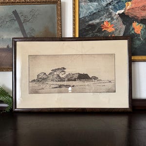 May include: A framed black and white etching depicting a rural landscape with a thatched-roof cottage and trees. The artwork is presented in a dark brown wooden frame with a cream-colored mat board and features a signature in the bottom right corner.
