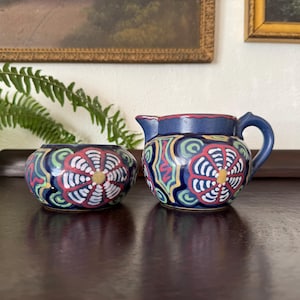 May include: A ceramic creamer and sugar bowl set. The creamer has a handle and spout, and the sugar bowl is round. Both pieces are navy blue with colorful floral patterns in red, white, yellow, and green. The set is on a dark brown surface.