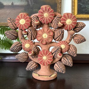 Pu&ograve; includere: Una scultura in terracotta a forma di albero della vita con cinque grandi fiori rossi dai centri gialli, circondati da numerose foglie marrone scuro con venature incise e piccoli boccioli rotondi su una base circolare.