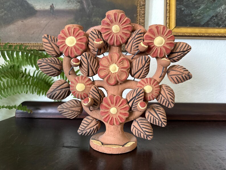 Pu&ograve; includere: Una scultura in terracotta a forma di albero della vita con cinque grandi fiori rossi dai centri gialli, circondati da numerose foglie marrone scuro con venature incise e piccoli boccioli rotondi su una base circolare.