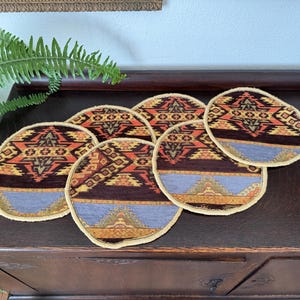 May include: Set of six round woven coasters with a geometric pattern in shades of brown, orange, yellow, and blue. The coasters have a cream-colored fringe around the edges.