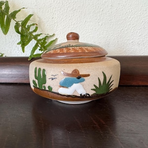 May include: A ceramic jar with a lid, featuring a hand-painted design of a person in a sombrero, cactus, and desert landscape. The jar is beige with brown and green accents, and the lid has a wooden knob.
