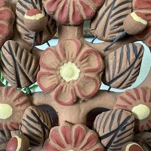Pu&ograve; includere: Una scultura decorativa in argilla con un fiore centrale dai petali color terracotta e centro giallo pallido, circondato da altri fiori, foglie strutturate con venature marrone scuro e piccoli boccioli rotondi su sottili steli di filo.