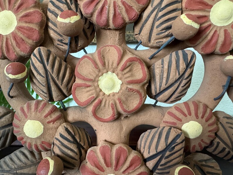 Pu&ograve; includere: Una scultura decorativa in argilla con un fiore centrale dai petali color terracotta e centro giallo pallido, circondato da altri fiori, foglie strutturate con venature marrone scuro e piccoli boccioli rotondi su sottili steli di filo.
