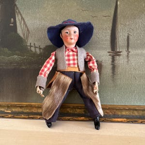 May include: A vintage doll dressed in a cowboy outfit. The doll has a painted face, a blue hat, a red and white plaid shirt, a gray vest, and faux fur chaps. The doll is standing in front of a landscape painting.