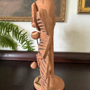 Pu&ograve; includere: Un portacandele in terracotta con un corpo cilindrico testurizzato decorato con sporgenze scolpite a forma di foglia. Le foglie sono decorate con motivi a strisce marrone scuro e piccole sfere rotonde multicolori sono fissate da sottili fili metallici.