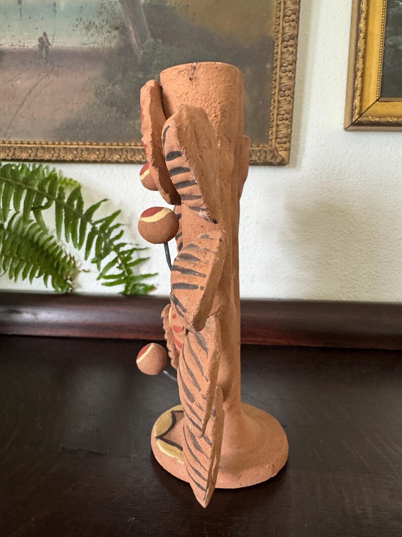 Pu&ograve; includere: Un portacandele in terracotta con un corpo cilindrico testurizzato decorato con sporgenze scolpite a forma di foglia. Le foglie sono decorate con motivi a strisce marrone scuro e piccole sfere rotonde multicolori sono fissate da sottili fili metallici.