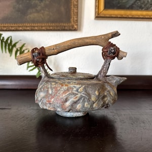 May include: A ceramic teapot with a textured, mottled grey and rust-colored surface featuring a raised leaf pattern. The teapot has a round lid and a handle made from a piece of natural driftwood, which is secured to the body with brown leather cords.