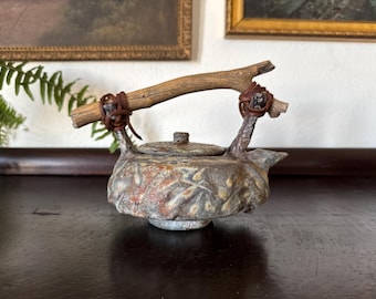 May include: A ceramic teapot with a textured, mottled grey and rust-colored surface featuring a raised leaf pattern. The teapot has a round lid and a handle made from a piece of natural driftwood, which is secured to the body with brown leather cords.