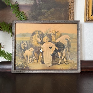 May include: Framed vintage print depicting a child surrounded by a lion, wolf, lamb, and cow. The artwork features a pastoral scene with soft colors and a silver-toned frame. The print evokes a sense of peace and harmony.