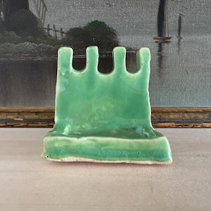 May include: A green ceramic spoon rest with a unique, castle-like design. The spoon rest has a rectangular base and a top with four arched cutouts. The ceramic has a glossy finish and a light green color.