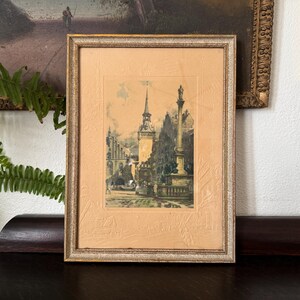 May include: A framed print featuring a detailed architectural scene of a European town square with a tall central spire and a monument column. The artwork is set within a textured, light peach-colored mat and a slim, weathered gold-toned frame.