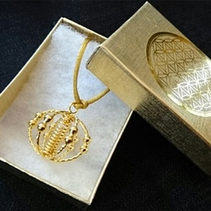 Könnte beinhalten: Eine goldene Halskette mit einem kugelförmigen Anhänger, der mit einem drahtgewickelten Design und kleinen goldenen Perlen verziert ist. Die Halskette wird in einer goldenen Schachtel mit einem geometrischen Muster präsentiert.