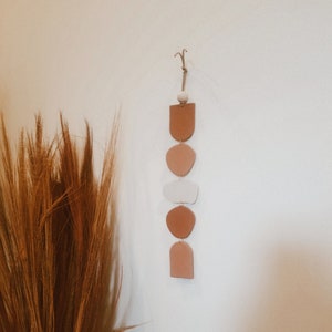 May include: A wall hanging made of five ceramic shapes in shades of brown, beige, and white. The shapes are suspended from a string and are arranged in a vertical line.