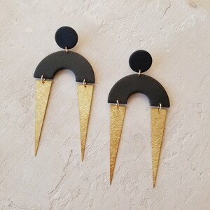 May include: A pair of black and gold geometric earrings. The earrings feature a black arch shape with a gold triangle on each side. The earrings are suspended from a black circle.