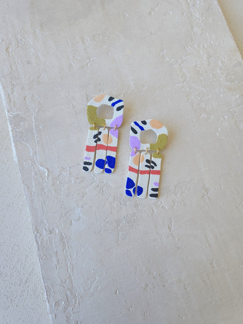 K&ouml;nnte beinhalten: Ein Paar H&auml;ngeohrringe mit geometrischem Design. Die Ohrringe sind aus wei&szlig;em Polymerton gefertigt und weisen eine Vielzahl von Farben auf, darunter Blau, Gr&uuml;n, Pink und Orange. Die Ohrringe sind quadratisch geformt mit einem kleineren Quadrat, das aus der Mitte herausgeschnitten ist. Die Ohrringe sind an einem goldenen Haken befestigt.