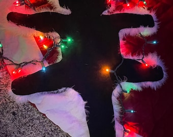 May include: A black cat-shaped Christmas decoration with white trim, lying on a red and white surface. Colorful Christmas lights are strung around the cat. The decoration is on a textured, gray surface.