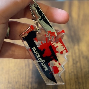 May include: A clear acrylic keychain with a black knife design and red blood splatter. The knife has the text "Slice of Life" printed on it.