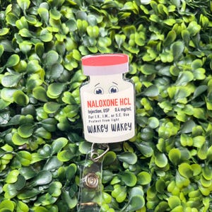 May include: A badge reel featuring a white vial with a red cap and a cartoon face. The label reads NALOXONE HCL, Injection, USP 0.4 mg/mL, For I.V., I.M., or S.C. Use, Protect from light, and WAKEY WAKEY. The vial is attached to a clear plastic clip.
