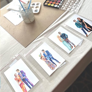 May include: Watercolor illustrations of couples in various outfits, displayed in clear plastic sleeves. A jar with paintbrushes, a watercolor palette, and art supplies are on a table. The artwork features vibrant colors and detailed clothing.