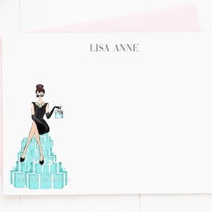 May include: A personalized stationery set featuring a woman in a black dress and sunglasses sitting on a pile of turquoise gift boxes. The text "LISA ANNE" is printed at the top of the card.