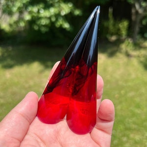 Puede incluir: Un par de pendientes de resina con un diseño degradado. Los pendientes hacen la transición de un rojo vibrante en la base a un negro profundo en las puntas puntiagudas. Los pendientes se sostienen en una mano sobre un fondo verde borroso.