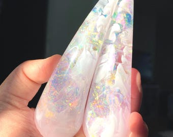 May include: Two clear, teardrop-shaped resin objects with iridescent glitter and white floral inclusions. The objects have a soft pink base and are held in a hand, with a dark background.