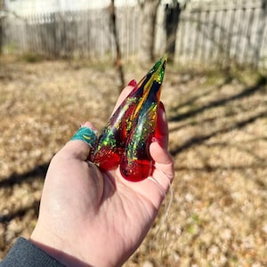 Könnte beinhalten: Zwei lange, spitze Nagelspitzen aus Kunstharz. Die Spitzen haben einen Farbverlauf aus Rot, Gold, Grün und Schwarz mit Glitzerakzenten. An einer der Nagelspitzen ist ein dünner, klarer Faden befestigt.