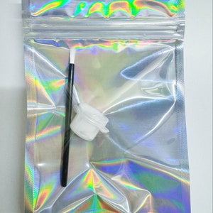 Puede incluir: Una bolsa transparente iridiscente con cierre zip que contiene un pincel con mango negro y un pequeño recipiente de pintura blanco con tapa. La bolsa refleja un arcoíris de colores. Para proyectos de arte o manualidades.