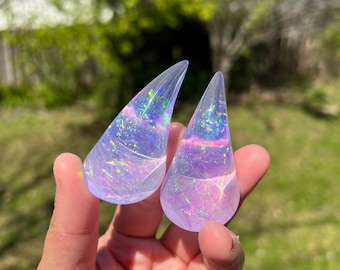 May include: Two iridescent, teardrop-shaped resin objects with a swirling, colorful interior. The objects are held in a hand against a blurred green background. The resin has a translucent quality, catching light and displaying a range of colors.