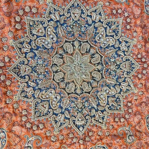 May include: A close-up of a colorful fabric with a large, intricate, embroidered design in the center. The design is in shades of blue, gold, and white, and features a floral motif. The fabric is a vibrant orange color with a paisley pattern.