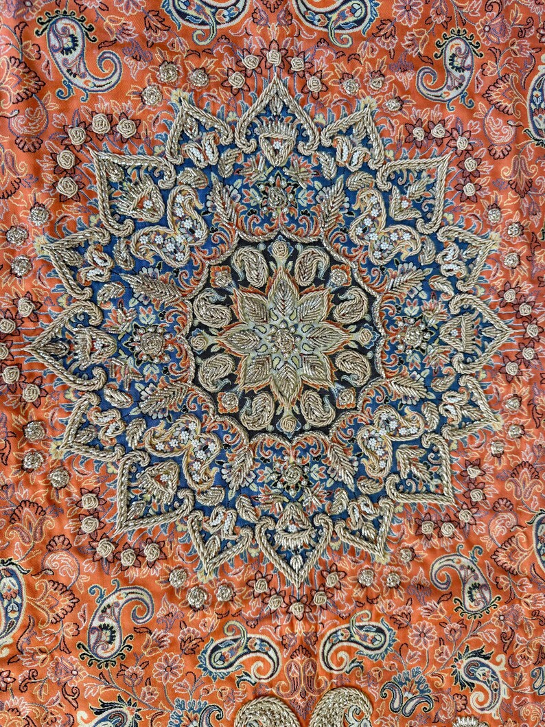 May include: A close-up of a colorful fabric with a large, intricate, embroidered design in the center. The design is in shades of blue, gold, and white, and features a floral motif. The fabric is a vibrant orange color with a paisley pattern.