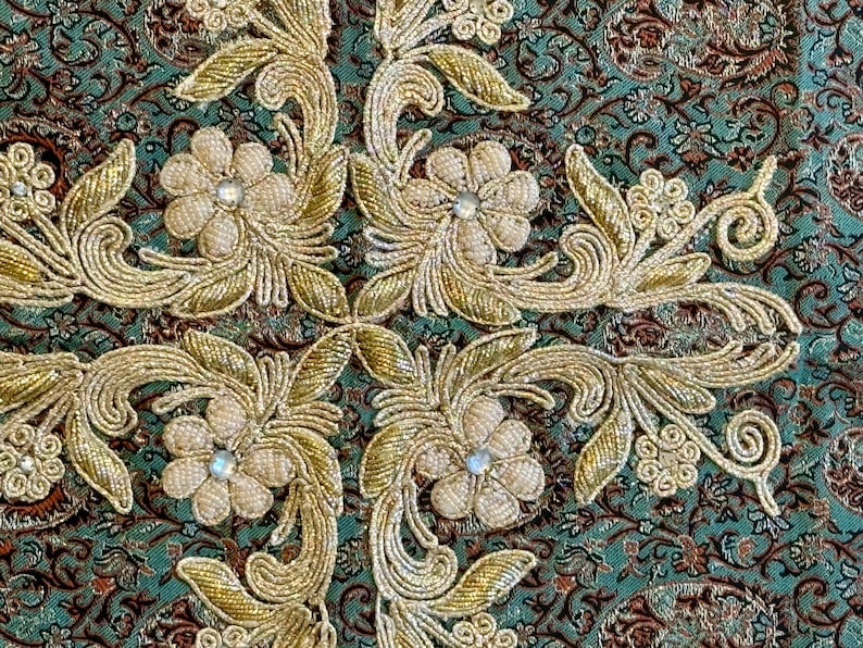 May include: Close-up of a teal fabric with gold embroidery of floral designs. The fabric has a repeating pattern of small flowers and leaves.