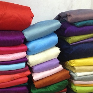 Puede incluir: Una pila de rollos de tela de colores en varios tonos de azul, morado, rosa, rojo, amarillo, verde y marrón. La tela está doblada cuidadosamente y dispuesta en una fila.