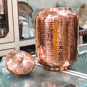 May include: A cylindrical copper water dispenser featuring a hammered texture, a removable lid with a small knob, and a silver-toned metal spigot attached to the lower side. The vessel has a polished finish that reflects light across its surface.
