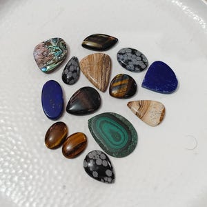 Puede incluir: Una colección de piedras preciosas pulidas de varias formas y colores. Incluye un lapislázuli azul, una malaquita verde y piedras ojo de tigre. Las piedras preciosas se exhiben sobre una superficie blanca.