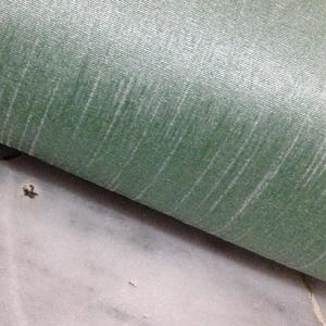 Puede incluir: Primer plano de una sección doblada de tela verde con vetas verticales blancas. La tela tiene un brillo sutil y una apariencia texturizada. El fondo es una superficie clara, posiblemente de mármol.