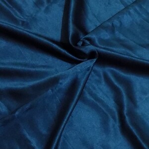 May include: Close-up of a folded piece of teal-colored satin fabric. The fabric has a smooth, shiny texture with visible folds and creases, creating shadows and highlights. The color is a deep, rich teal, with a slight sheen.