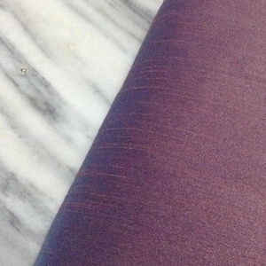 Puede incluir: Primer plano de una sección de tela morada con un brillo sutil, en ángulo contra una superficie de mármol blanco y gris. La tela tiene una apariencia texturizada con fibras visibles y un degradado de color.