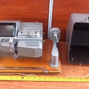 Puede incluir: Una máquina de edición de películas plateada vintage con una pantalla negra y varias perillas y palancas. Se encuentra sobre una base de madera con un asa plateada a cada lado. Una caja gris está a la derecha. Una cinta métrica amarilla está debajo.