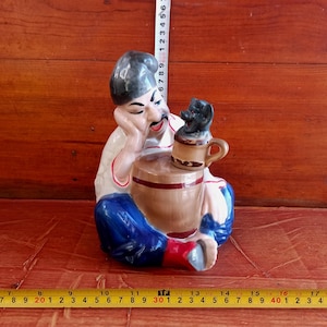 Könnte beinhalten: Keramikfigur eines Mannes in traditioneller Kleidung, der sich auf ein Fass stützt. Die Figur ist mit einem grauen Hut, einem weißen Hemd, einer blauen Hose und roten Stiefeln bemalt. Ein kleiner Hund sitzt auf einem Becher. Höhe ca. 15 cm.