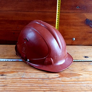 Puede incluir: Un casco de seguridad marrón con una etiqueta blanca que dice "TPL" en el lateral. El casco está sobre una superficie de madera.