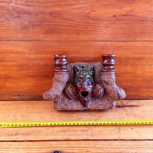 May include: A ceramic wall sconce with a brown and green glazed finish. The sconce features a grotesque face with a wide open mouth and two candle holders on either side.