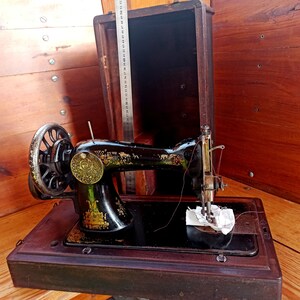 May include: A vintage black sewing machine with a wooden case. The machine has a metal foot pedal and a needle threader. The case is made of dark wood and has a hinged lid.