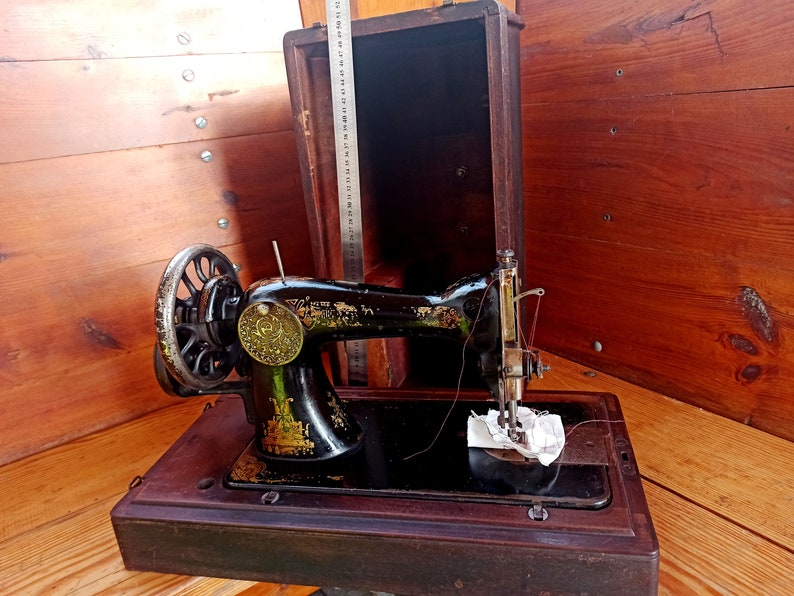 May include: A vintage black sewing machine with a wooden case. The machine has a metal foot pedal and a needle threader. The case is made of dark wood and has a hinged lid.